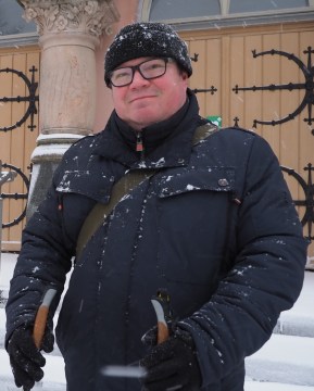 Silmälasipäinen, tummiin vaatteisiin pukeutunut mies seisoo kirkon portailla.