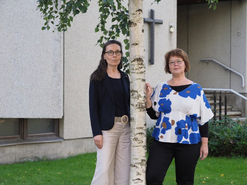 Kaksi naista seisoo koivun juurella seurakuntatalon edessä.
