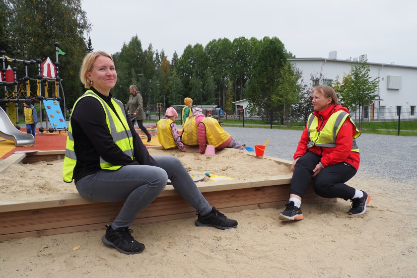 Kaksi lastenhoitaa istuu hiekkalaatikolla lasten leikkiessä.