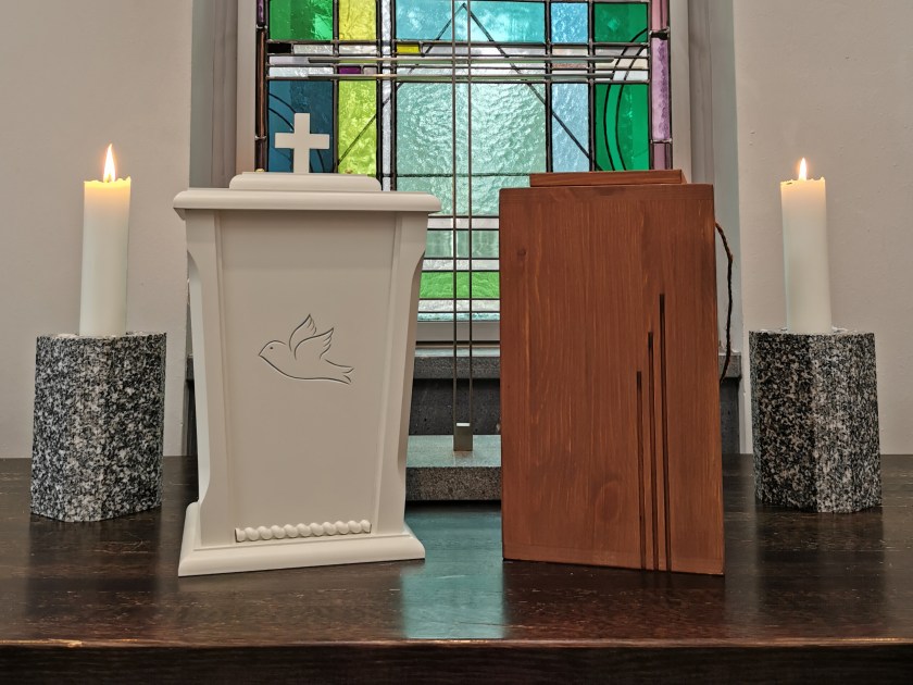 Valkoinen ja puunvärinen tuhkauurna, taustalla näkyy lkirkollinen asimaalaus.