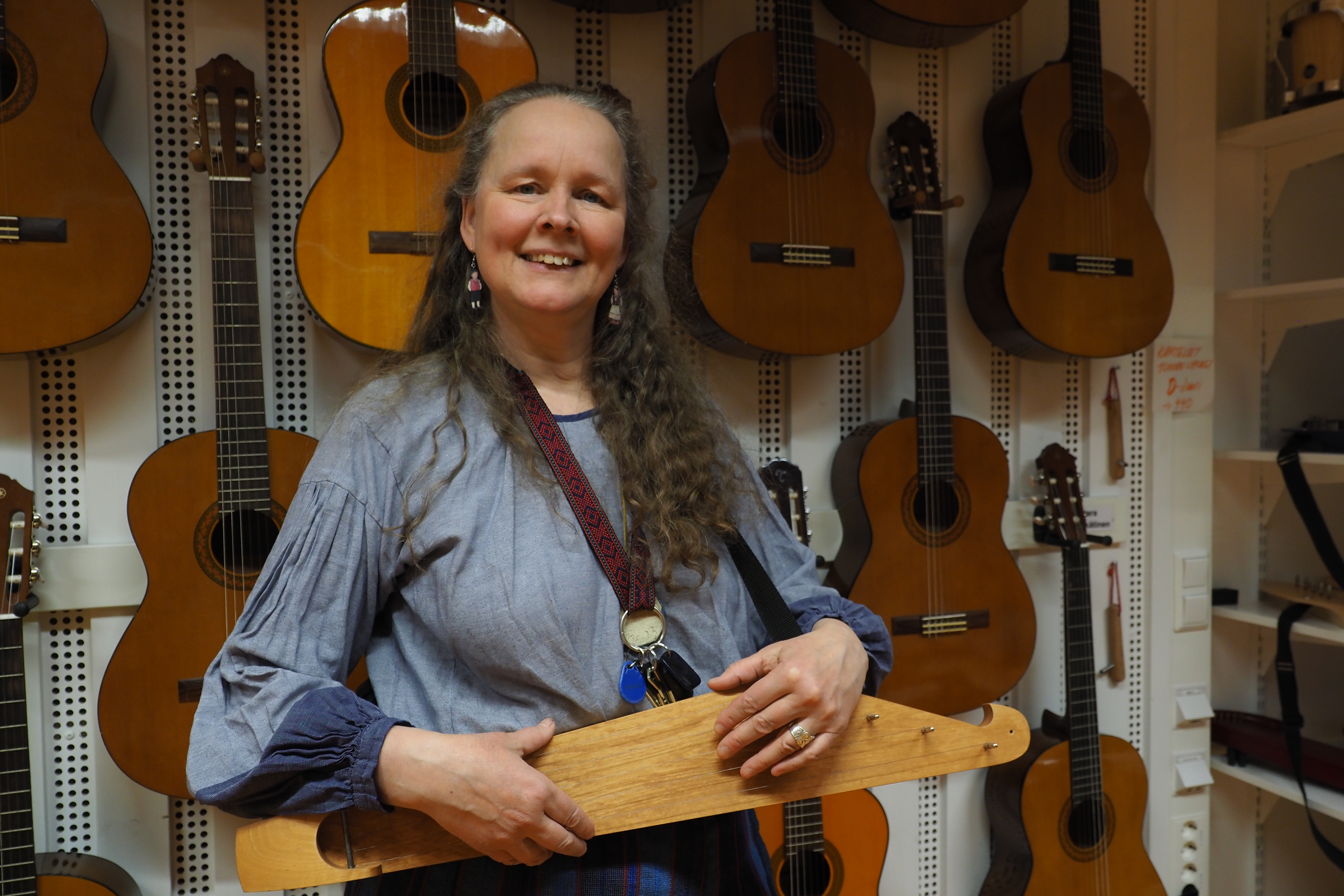 Pitkähiuksinen nainen kanteleen kanssa, takana seinällä kitaroita.