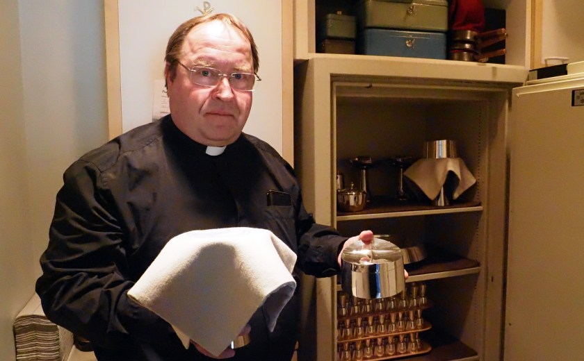 Kirkkoherra Ari Autio seisoo ehtoollisvälineitä sisältävä kaapin edessä ehtoollismalja ja ehtoollileipäastia kädessään. 