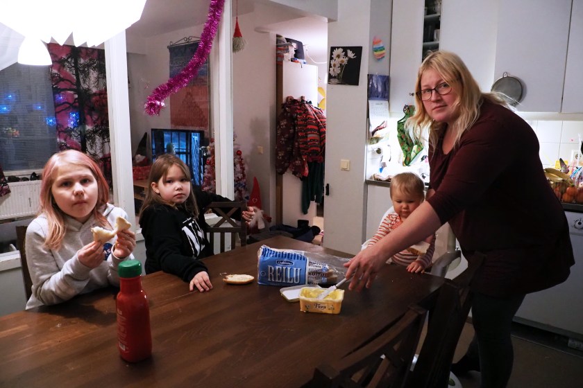 Kolme lasta istuu pöydän ääressä syömässä paahtoleipää. Äiti Niina Väätäinen kattaa pöytää ja katsoo kameraan.