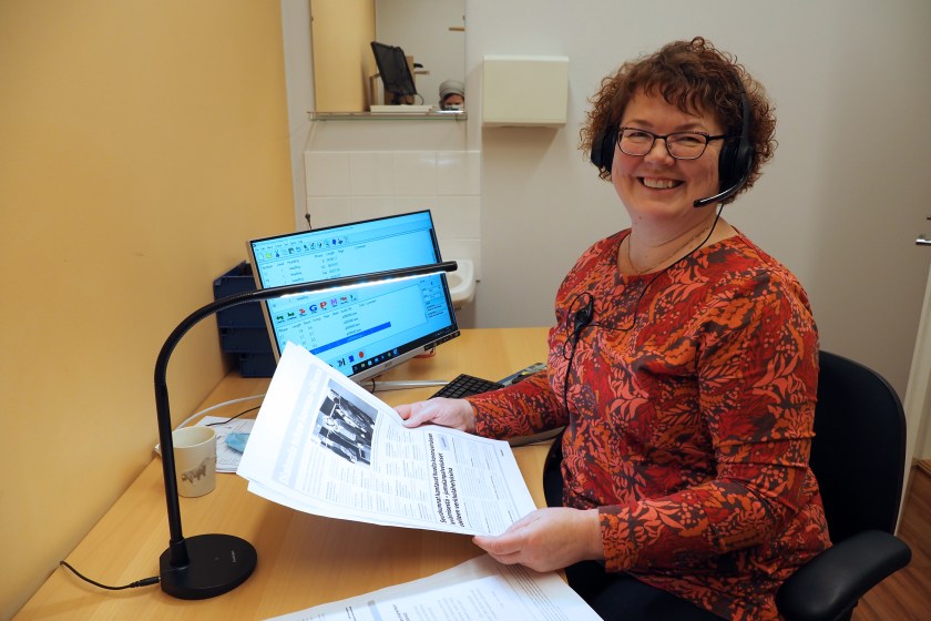 Ilona Suojama istuu työpöydän ääressä Kirkkotie-lehden sivut kädessään ja mikrofinikuulokkeet päässään, katsoo kameraan ja hymyilee. Taustalla tietokone, jossa käynnissä äänilehden äänitykseen tarvittava ohjelma.