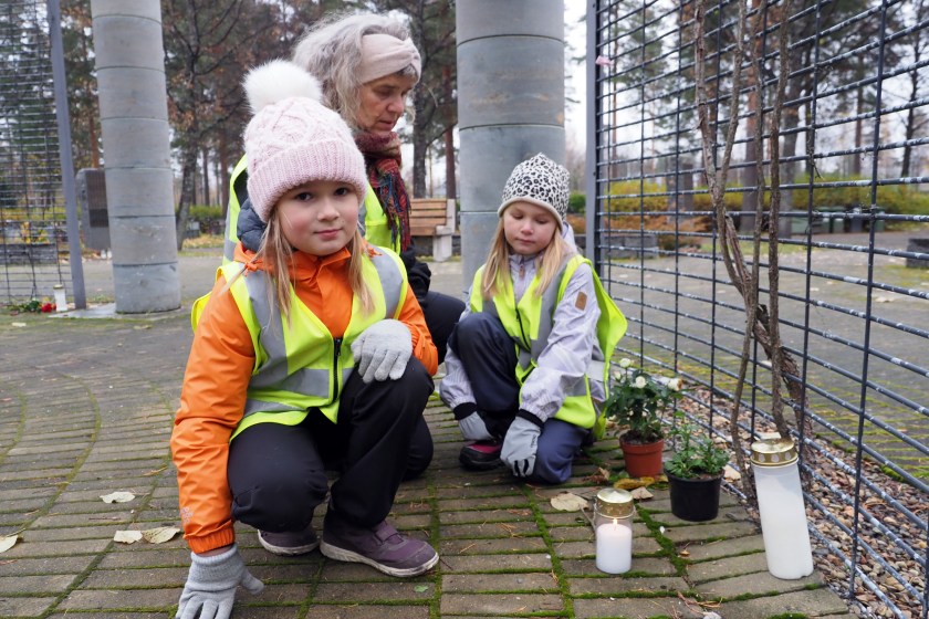 Kaksi lasta kyykistyneenä kynttilän ääreen yhdessä lastenohjaajan kanssa hautausmaan muistelupaikalla.