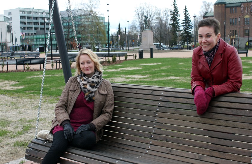 Diakoni Katja Nuuhkarinen istuu keinussa Joensuun torin laidalla ja pastori Kaisa Puustinen nojailee hymyillen keinun selkämykseen.