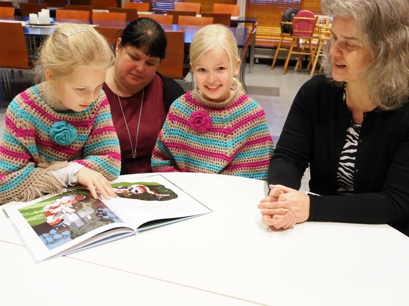 Iltapäiväkerholaiset ja lastenohjaajat istuvat pöydän ääressä lukemassa kirjaa.